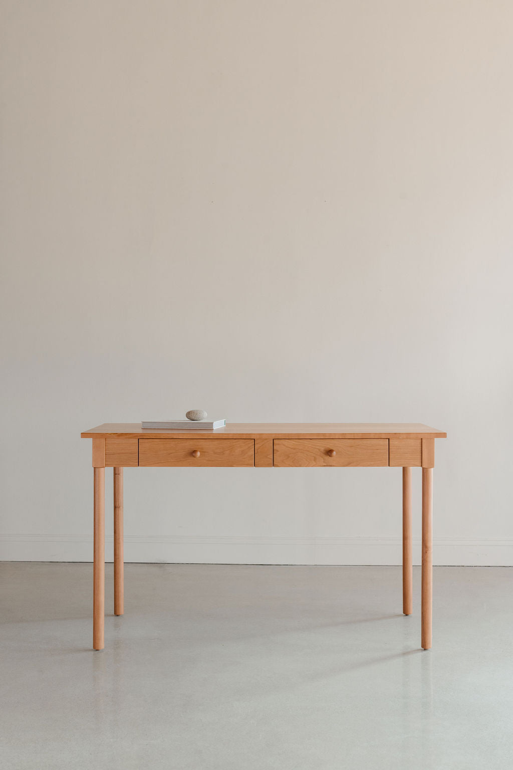 Writing desk in clean bright room with round legs and two drawers, and topped with a book and a rock